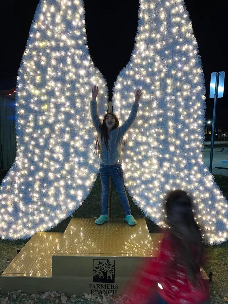 Alexa with arms raised in joy under a lit angel-wings display at night